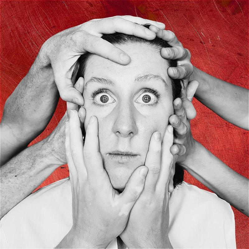 A black-and-white photo of a stunned person who stares ahead as six hands hold their head in place, beyond a red background.