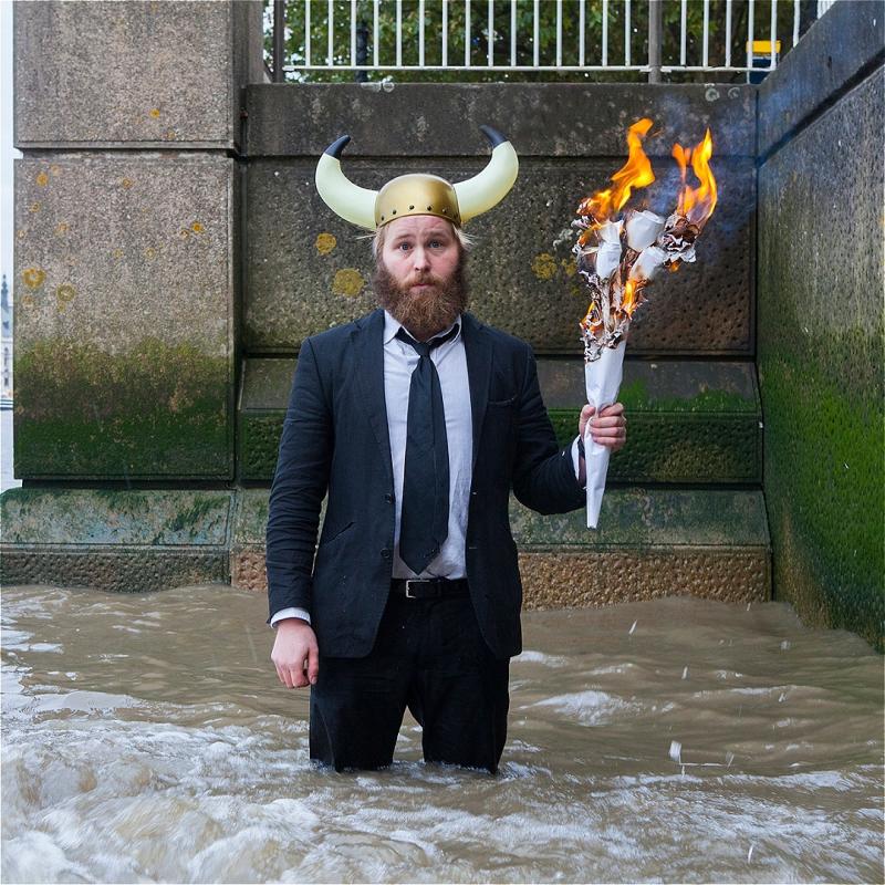 James stands knee-deep in a river, wearing a suit and a cartoony viking helmet. He holds a flaming torch while looking sadly at the camera.