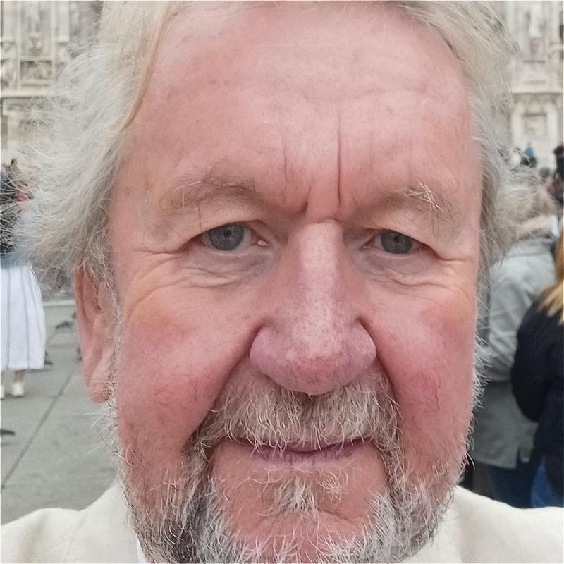 Close up of Pip's face as he stares ahead, smiling, in a public space with people behind him.