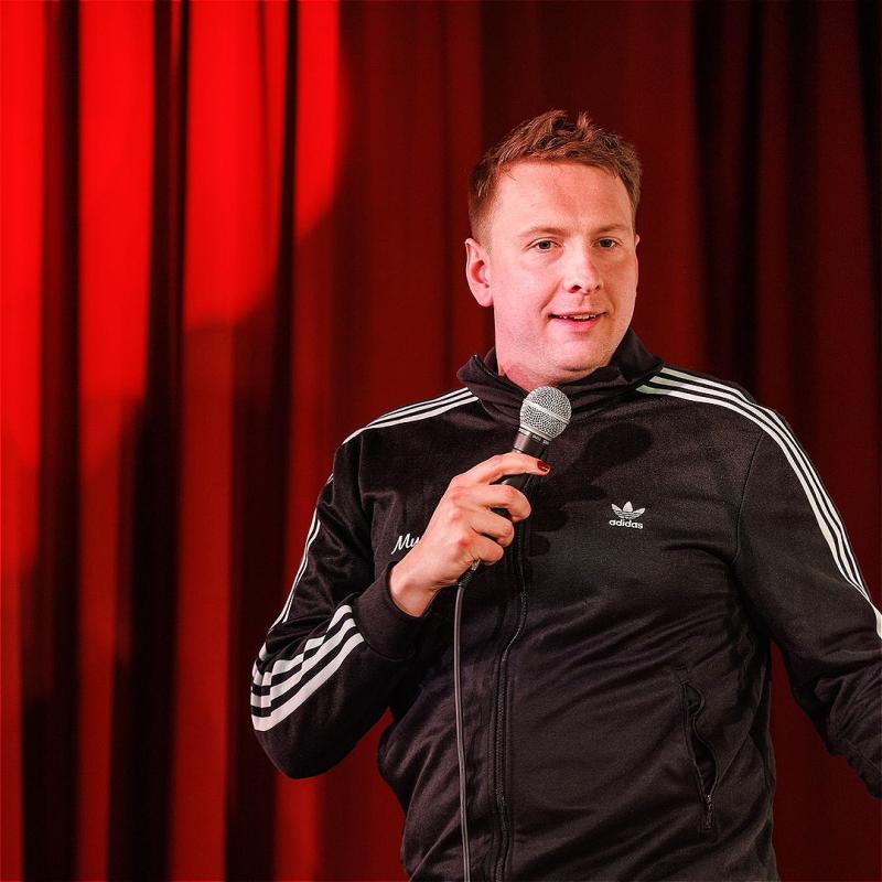 Joe Lycett wearing a black Adidas jacket and holding a microphone in front of a red curtain.