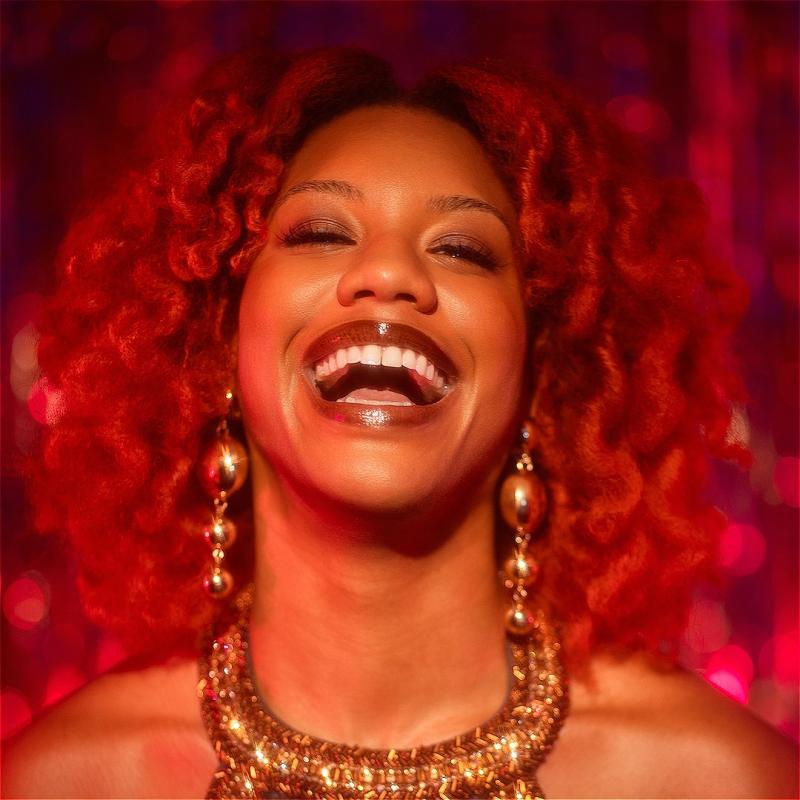 Kemah is wearing a gold sparkly halter top with matching earrings with a big laughing smile on their face. Their hair is red and matches the background.