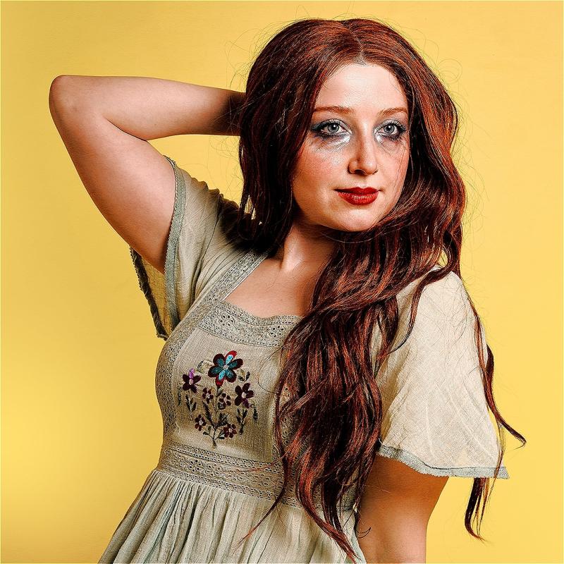 A woman with smudged makeup and wearing an old nightgown poses in front of a yellow background.