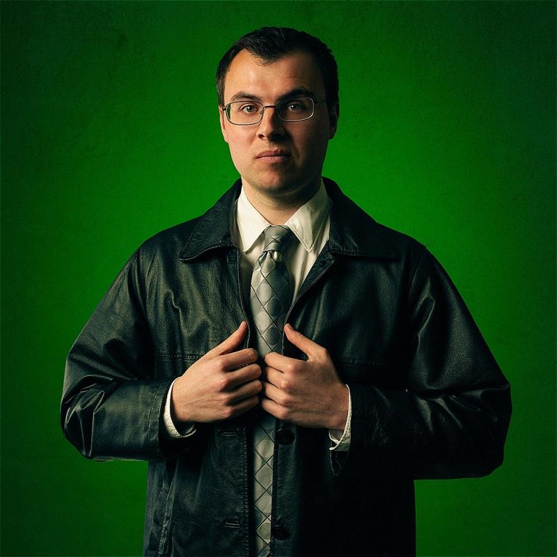 Freddie wears a leather jacket and a tie, staring directly at the camera, before a forest green background.