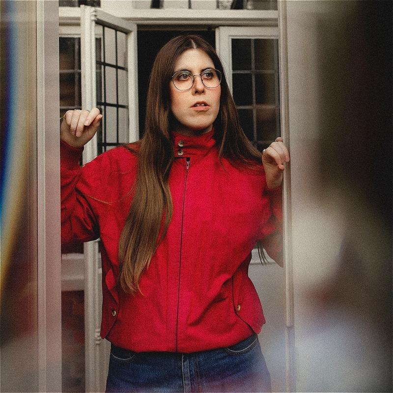 Ele, wearing a red jacket and jeans, stares out of a house, windows behind her.