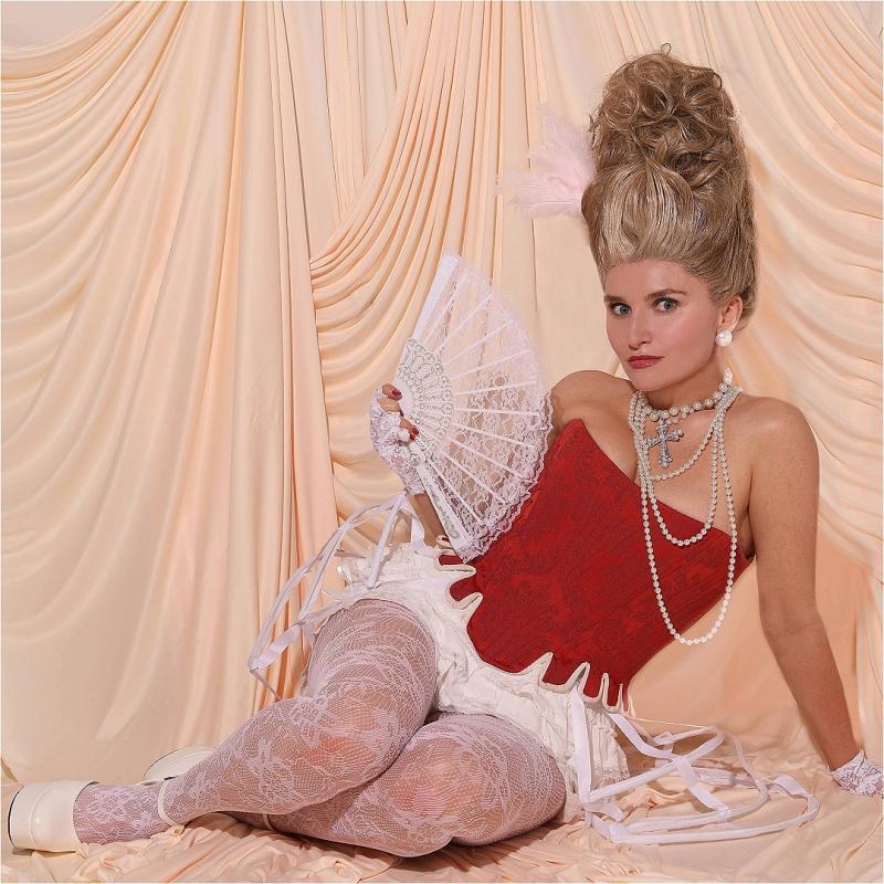 Sophie sitting down in a Marie Antoinette style outfit with an elaborate updo, red corset , pearls and a white lace fan. 