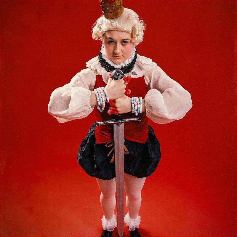 Lil standing holding a comically large sword and a small crown perched on their head like a petulant child.