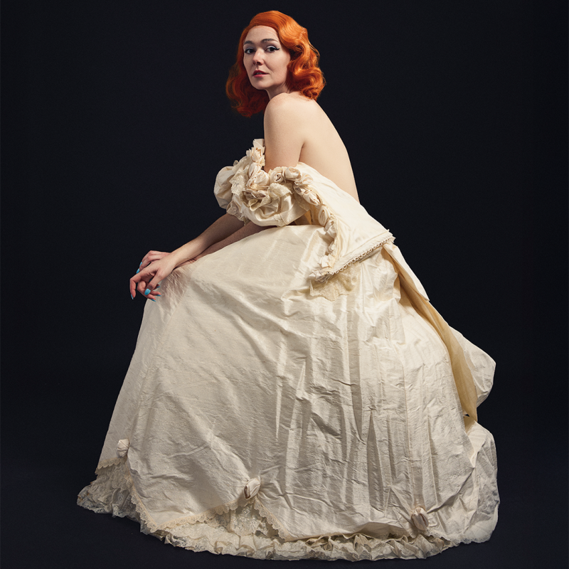 Elf sitting demurely in a unzipped floor length ballgown against a black backdrop.
