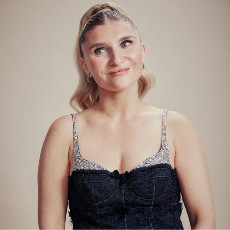 Sophie Garrad is standing against a pink background wearing a sparkly top. She's got her hair up in a slick ponytail and is smiling, looking off to the left.