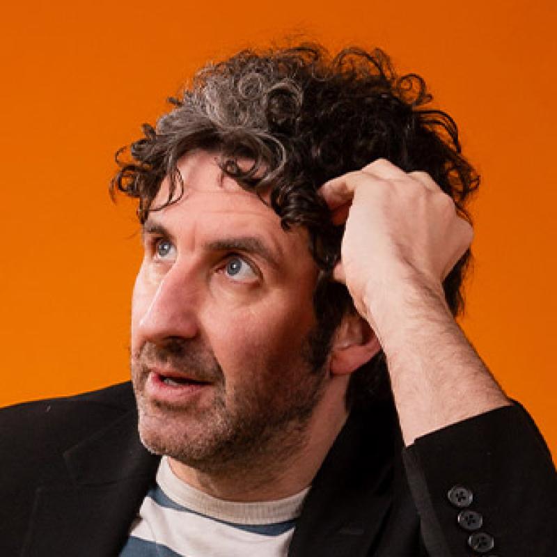 Mark Watson, against an orange background, looking off to the left with his head resting on his left hand 