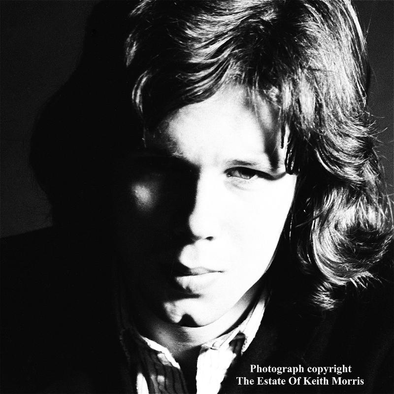 Black and white photo of a young man with shoulder-length hair staring straight ahead. 