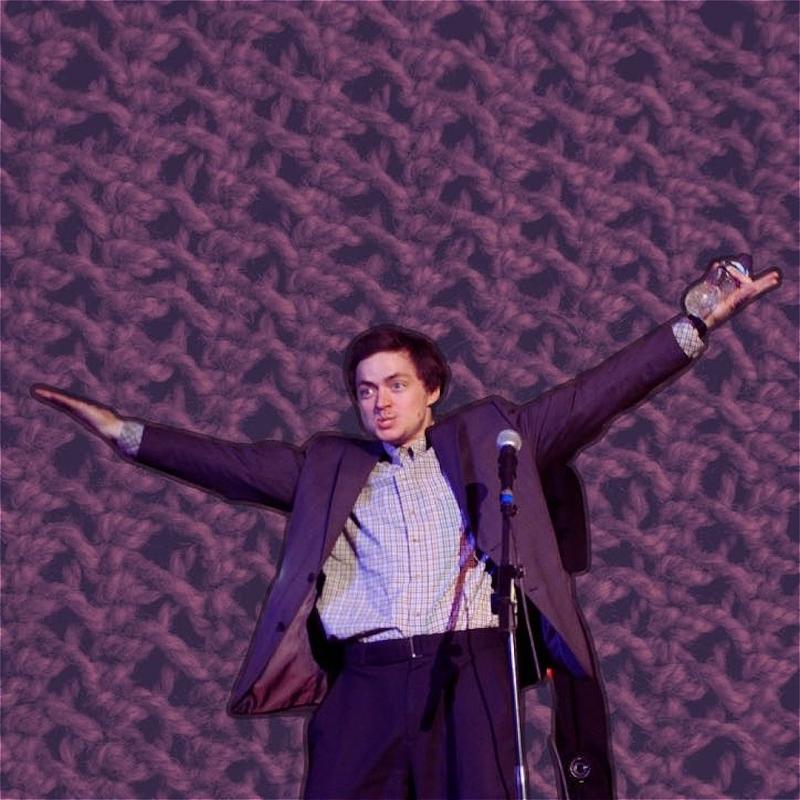 Tom is standing in front of a microphone, holding his arms out above him. He is wearing a check shirt and a purple suit jacket. The background is enlarged knit cloth in purple thread. 