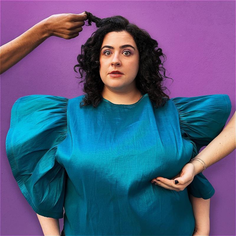 Jess Fuchs stands against a purple background, wearing a voluminous blue dress. Someone reaches into frame from the right and cups her breast. Someone else reaches in from the right and is holding a section of her black curly hair. 