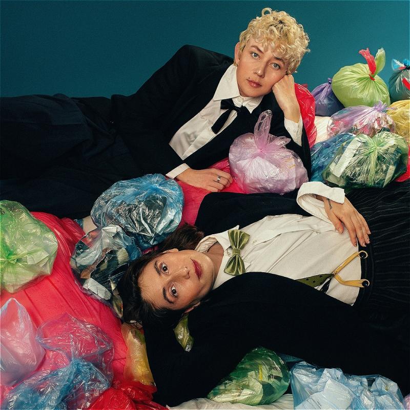 Two people in tuxedos are lying on top of a pile multicoloured bin bags