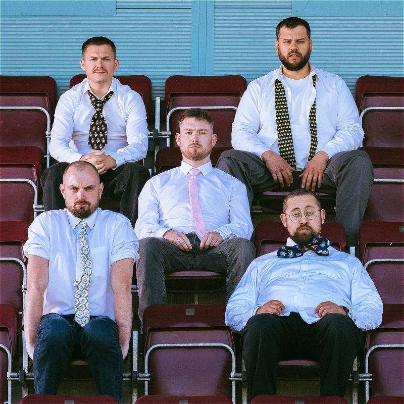 Five men sit in bleacher style seats, all wearing a shirt and tie and looking dishevelled. 