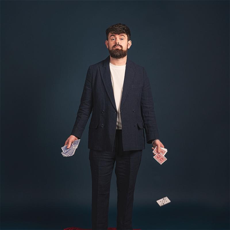 Tom in a black suit jacket, holding playing cards in both hands and one card is falling to the floor