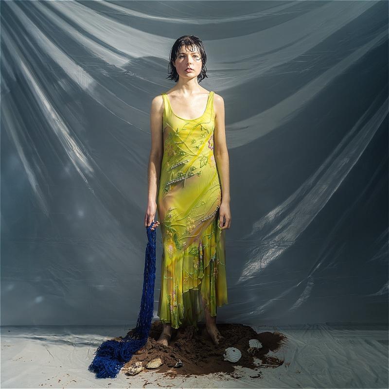 The performer stands in a sheer yellow/green dress and holding a blue fishing net trailing on the floor and standing in a small pile of mud.
