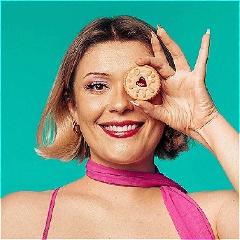 Cecily against a bright blue background, smiling and holding up a jammie dodger over one of her eyes. 