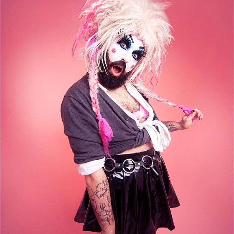 Baby Lame standing in front of a pink backdrop wearing Britney Spears' iconic schoolgirl outfit and a pink voluminous wig with pigtails. 
