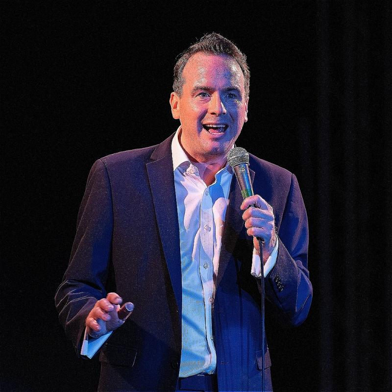 A stage picture of Matt Forde, a white man with brown hair wearing a white shirt and black suit jacket, speaking into a hand held mic, in front of a black background.