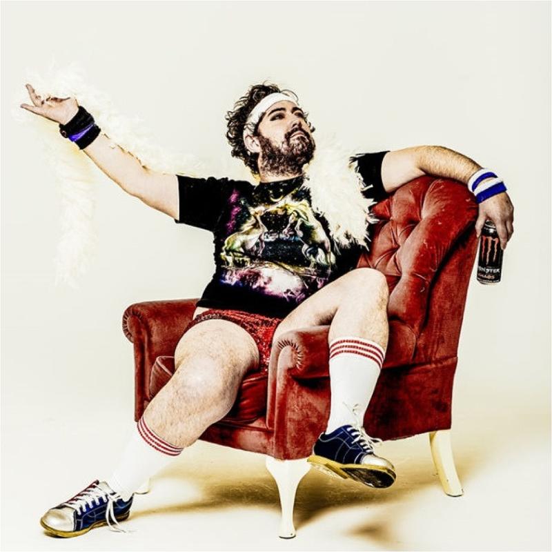 Nick Helm, a white man with brown hair and a beard is sitting on a red padded leather chair with one leg on the armrest. He is wearing a white boa, sports cuffs and headband, sports socks and Converse shoes, and holding a Monster can in his left hand. He is looking up with his arms open.
