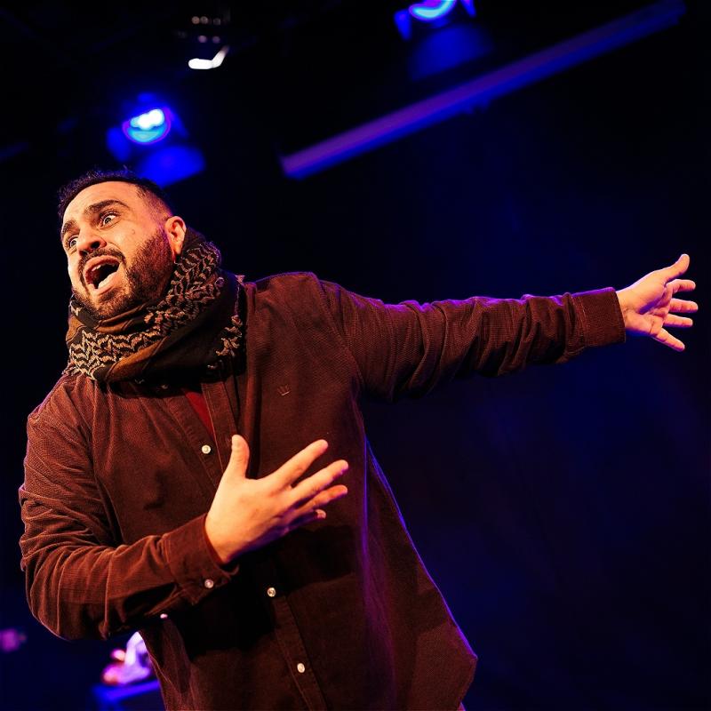 A stage mid shot of a brown man with a short beard, a keffiyeh around his neck and a burgundy shirt, looking out at the audience with an intense look and pointing at something behind him. There are blue spotlights in the background.