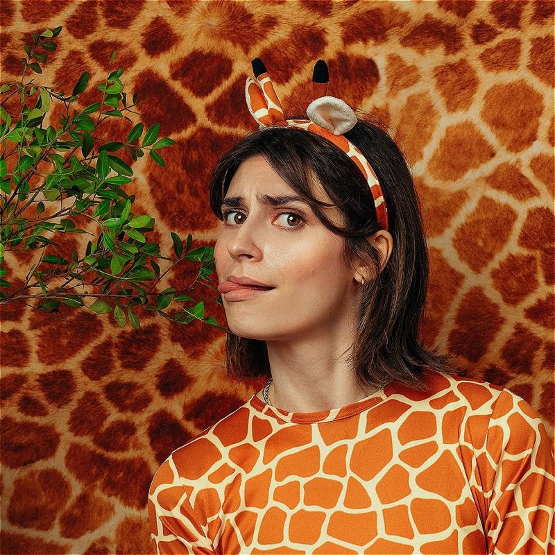 A woman wearing a giraffe patterned top and fake giraffe ears is licking a leaf on a branch that is coming into frame from the left. The background is giraffe pattern