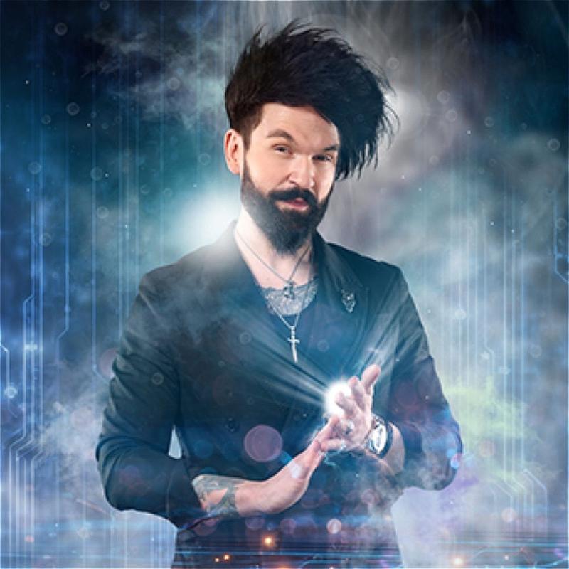 Colin Cloud, wearing a black suit jacket with chain necklaces, rubs his fingertips of one hand onto the palm of his other. A ball of light emenates from his palm. He is surrounded by smoke. 