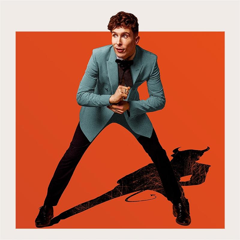 Larry is standing against an orange background, wearing a grey suit jacket and smart black trousers, leans over slightly and fixes a cufflink. Behind him his shadow casts down in the silhouette of a devil. 