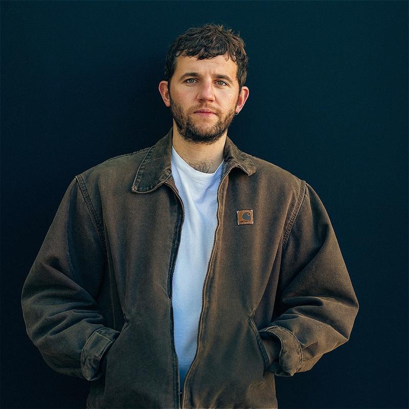 Rory leaning against a dark blue wall staring ahead into the camera wearing a Carhartt jacket.