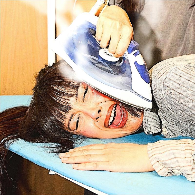 Patti Harrison's face is being pressed onto an ironing board by an unseen person, ironing her cheek with a blue and white iron. She has long brown hair and bangs and she is closing her eyes and opening her mouth as if she's screaming.