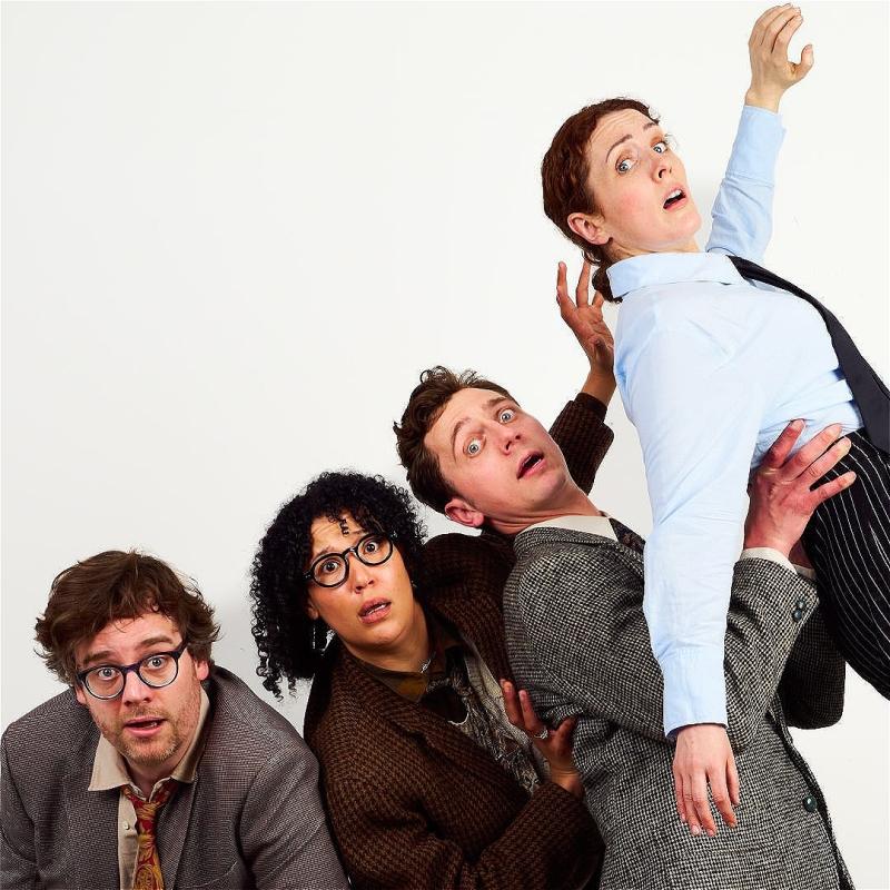 Cast of 4 performers dressed in suits and blazers looking alarmed against a plain white backdrop.