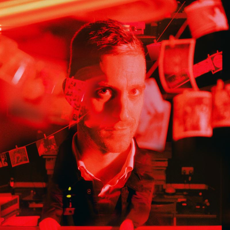Joseph in a dark room with Polaroids developing with a red tint over the image