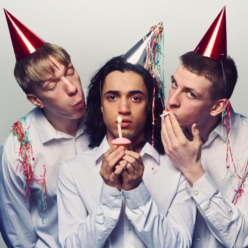 Three boys with party harts and white shirts. The middle boy holds a cupcake with a candle.