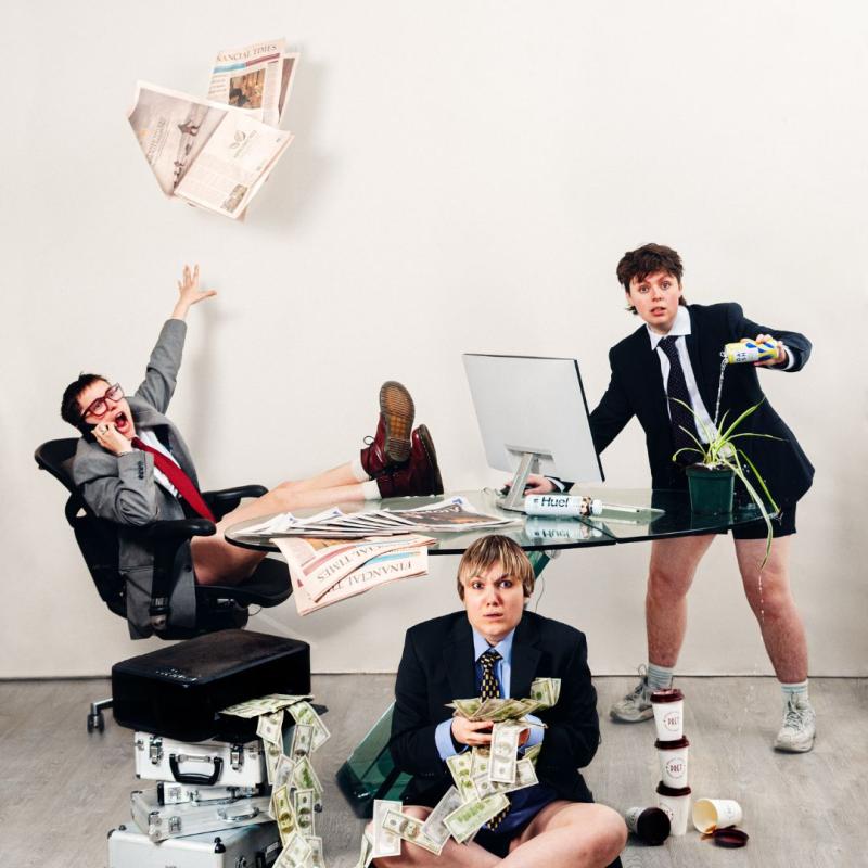 A chaotic scene of three people in suits around a glass desk. On the left is someone shouting on the phone and throwing a newspaper. On the right is someone in front of a computer, watering a plant with an energy drink. Sat on the floor, next to a pile of briefcases, is someone holding a large amount of cash 