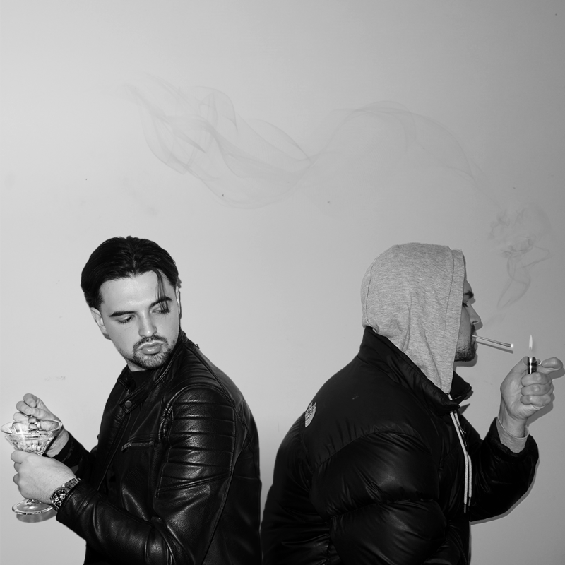Black and white image of two versions of the same man sitting back to back. One wears a leather jacket and holds a martini glass. The other wears a grey hoodie and puffer jacket with the hood up and is smoking.