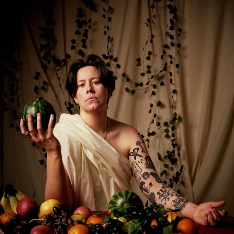 Chloe, holding a melon, wearing a toga sat in front of a table adorned with fruit. The curtain backdrop has leaves hanging in front. 