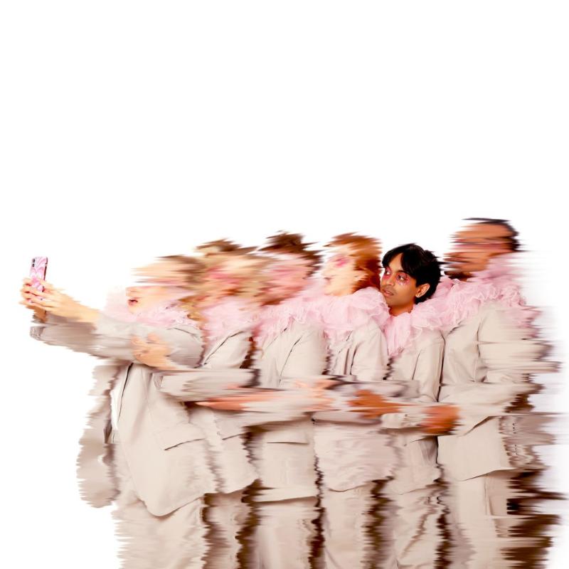 A blurry image of a line of performers standing one behind the other, wearing grey suits and pink ruffled collars. The person at the front is holding up a phone that they are all looking into 