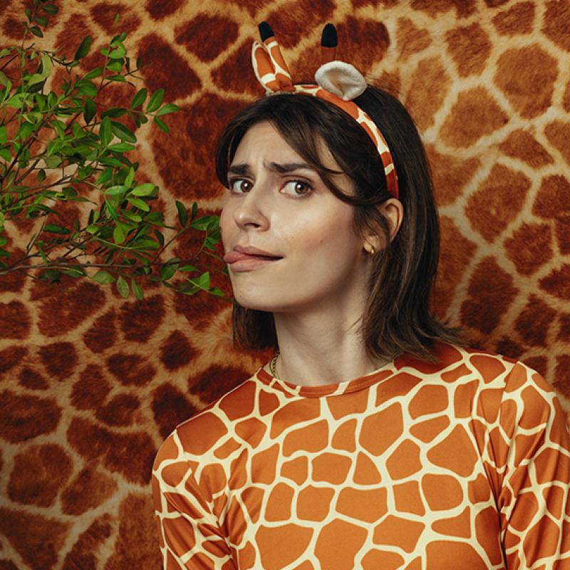 A woman wearing a giraffe patterned top and fake giraffe ears is licking a leaf on a branch that is coming into frame from the left. The background is giraffe pattern