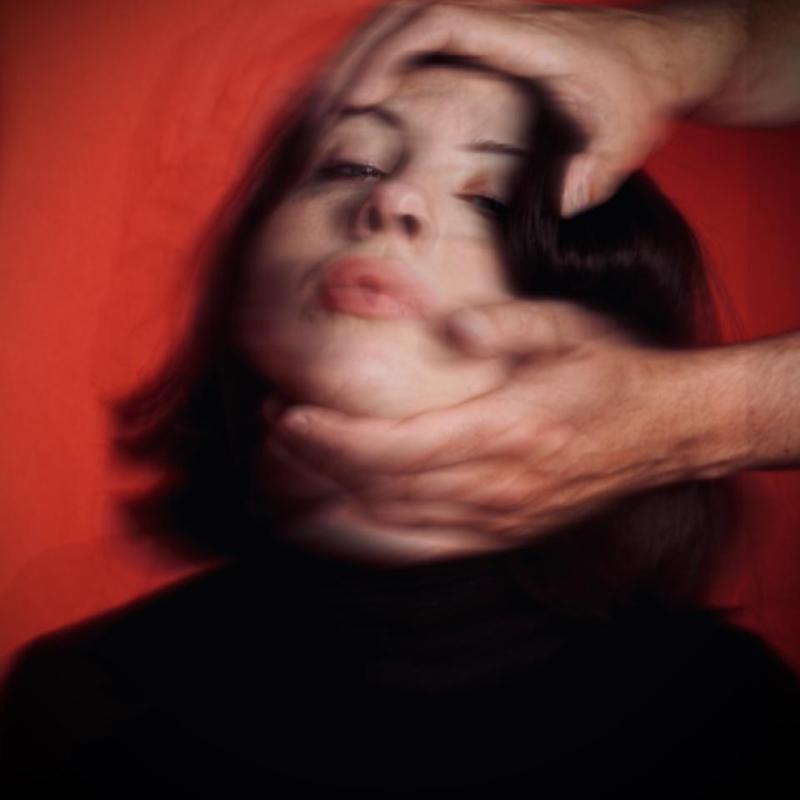 A blurry image of Harriet's face against a red background. Someone's hands reach in from out of frame and are holding the top of her head and her chin, pulling her head back so her face is tilted upwards