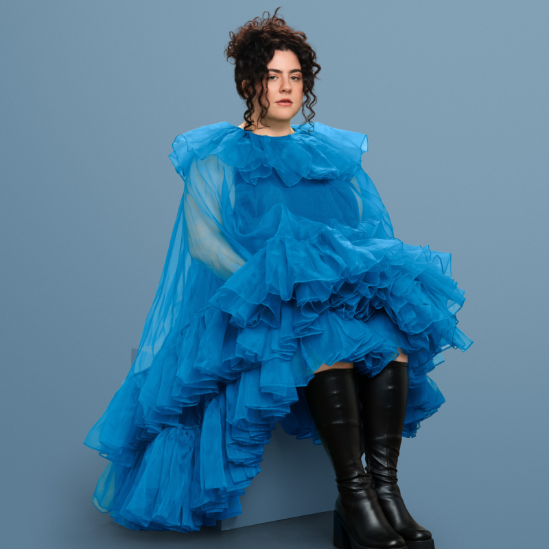 Jess wearing a blue tulle ruffled gown and a pair of leather knee-high boots sat on a block with with a vacant expression at the camera.