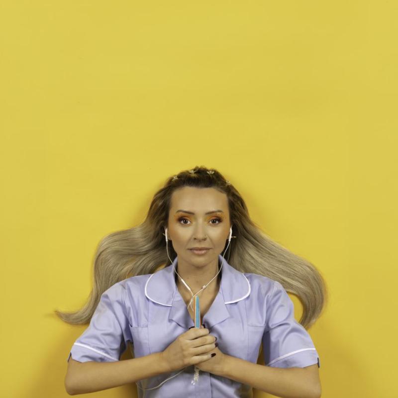 Kimberley is against a bright yellow background. They are wearing a blue nurses dress, with wired earphones in their ears. They are clasping a stick of rock between their hands, in front of their chest.  