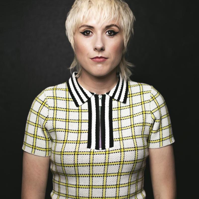 Maisie standing against a black background looking straight ahead. They are wearing a white t shirt with a yellow check pattern and black and white stripes on the collar