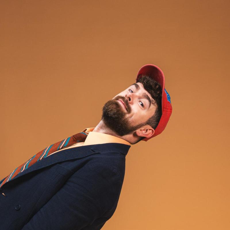 Tom brace dressed in a navy suit, orange shirt and tie and red baseball cap leaning back into frame agains an orange background