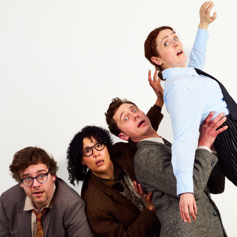 Cast of 4 performers dressed in suits and blazers looking alarmed against a plain white backdrop.