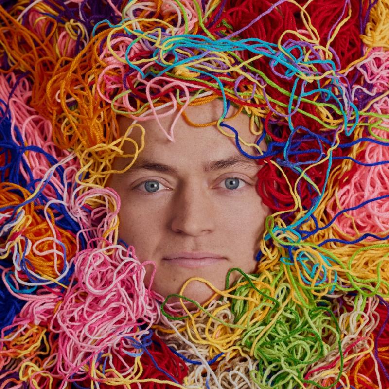 Tom's face is visible under a huge pile of different brightly coloured yarn 