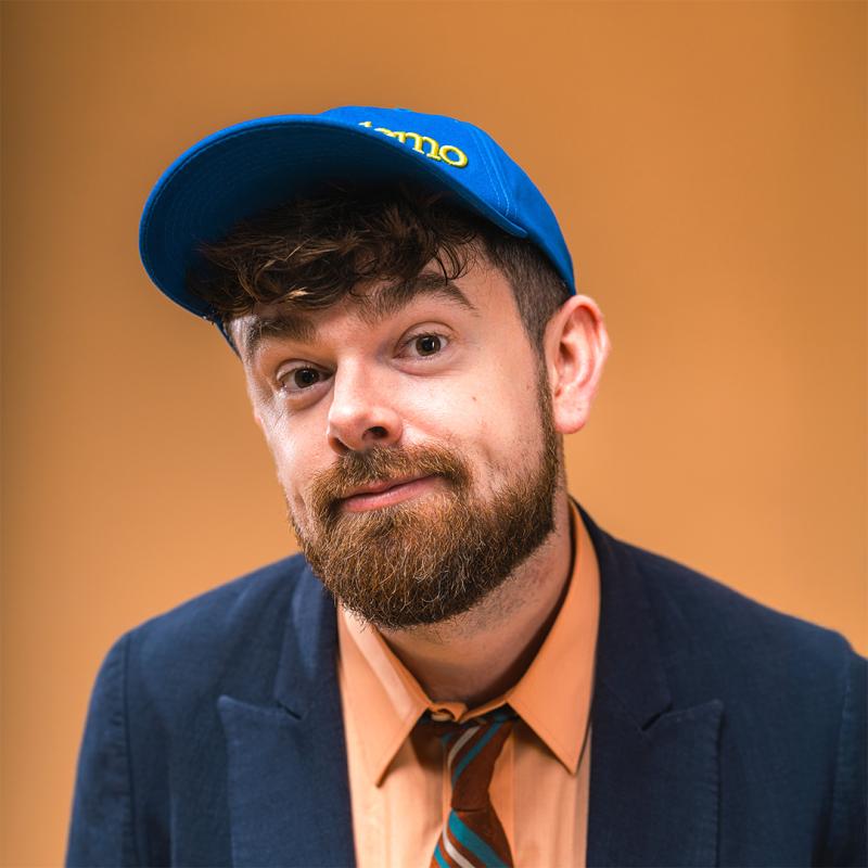 Tom brace dressed in a navy suit, orange shirt and tie and blue baseball cap on an orange background