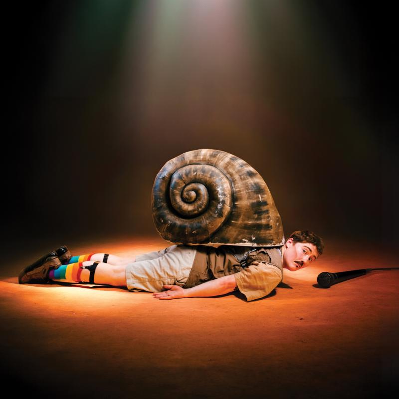 The performer lying flat with a large snail shell on their back. A microphone lies on the floor ahead of them.