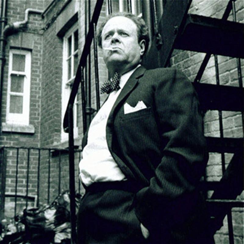 A Black and white image of a smartly dressed man leant against a metal staircase as a cigarette dangles from his mouth.