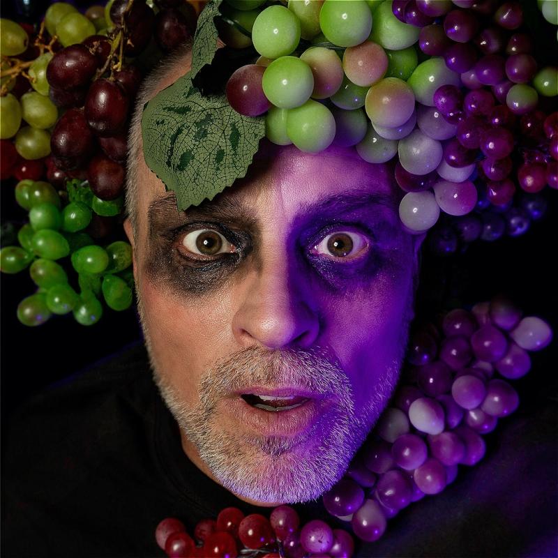 A man with a short white beard, brown eyes and black eyeshadow all around his eyes, is looking at the camera with bright eyes and his mouth slightly open. On his forehead and around his neck are white and red grapes and an ominous purple hue is lighting half of his face.