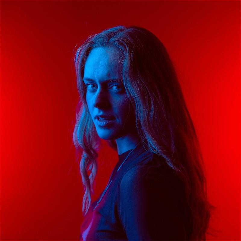A blonde person looks directly at the camera. They are submerged in a blue light, contrasting with the bright red background.